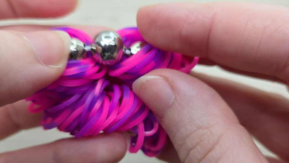 Hands holding a colorful pink and purple rubber band ball with silver beads, set against a blurred wooden background.