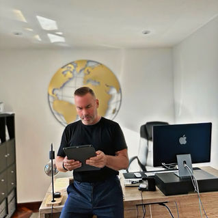 Eirik Moseng sitting on the edge of his desk, holding an iPad, with a world map on the wall and his workplace in the background, representing BEEJAYU’s mission to empower visually impaired individuals through technology.