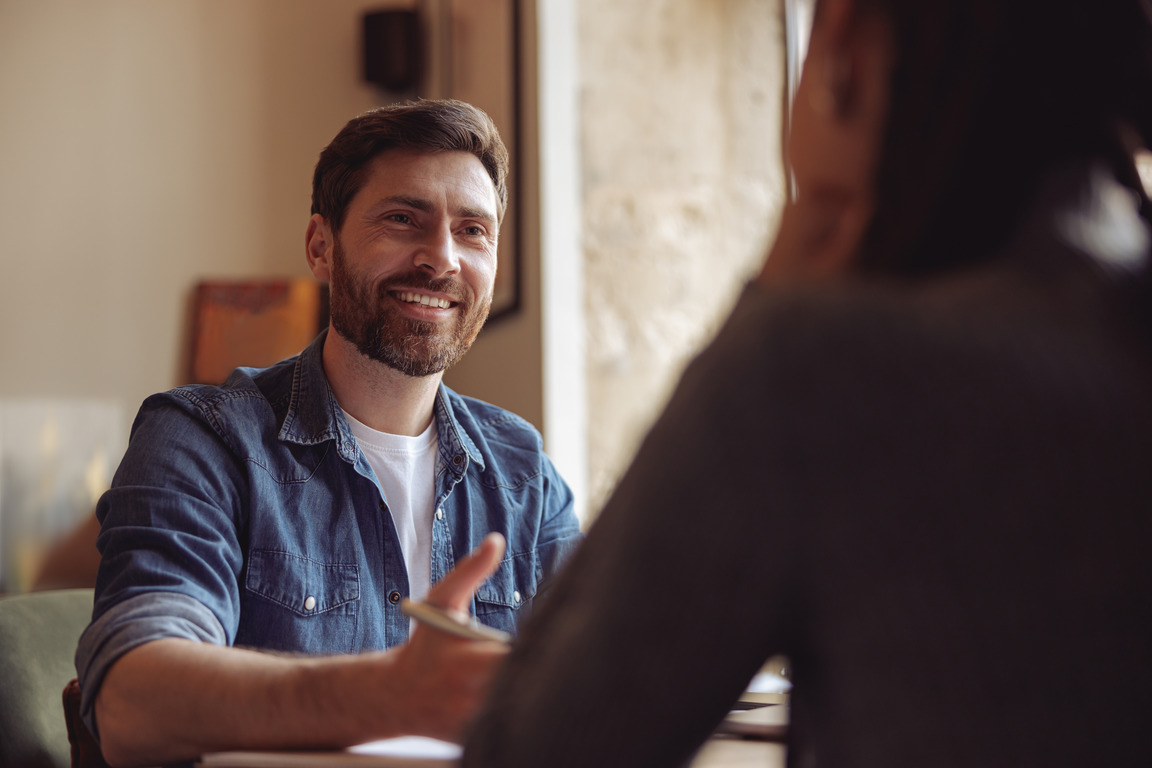 Smiling man talking, gesturing, casual setting, discussing ideas with another person.