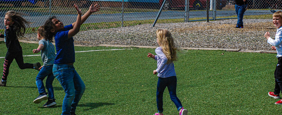 Five children are playing outside on the playground, four are running, one is reaching up to catch a football throw