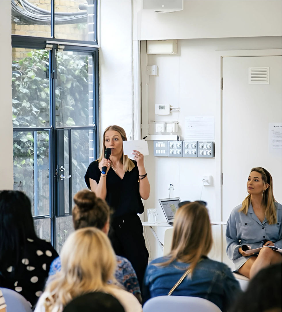 A woman in black is speaking and holding a card in a bright room with a large window. Seated audience listens, creating an attentive mood.