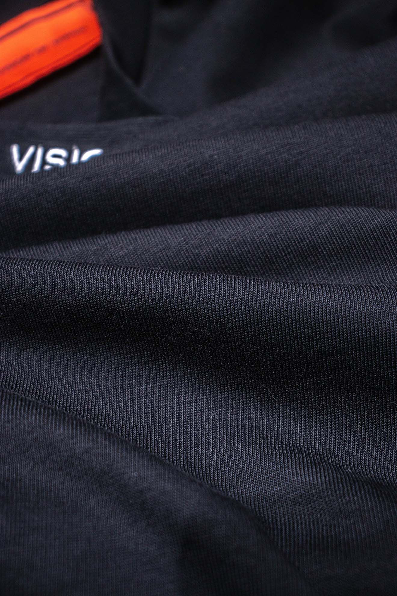 Áo khoác vải tối màu với chữ "VISION OF SUPER" thêu trắng, chi tiết cam.