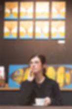 Portrait d’Audrey assise dans un café, regard tourné au loin avec un air contemplatif, lunette et main posée sur son visage. Elle semble réfléchie, comme si elle explorait une question intérieure importante. Une tasse de café chaud est devant elle.