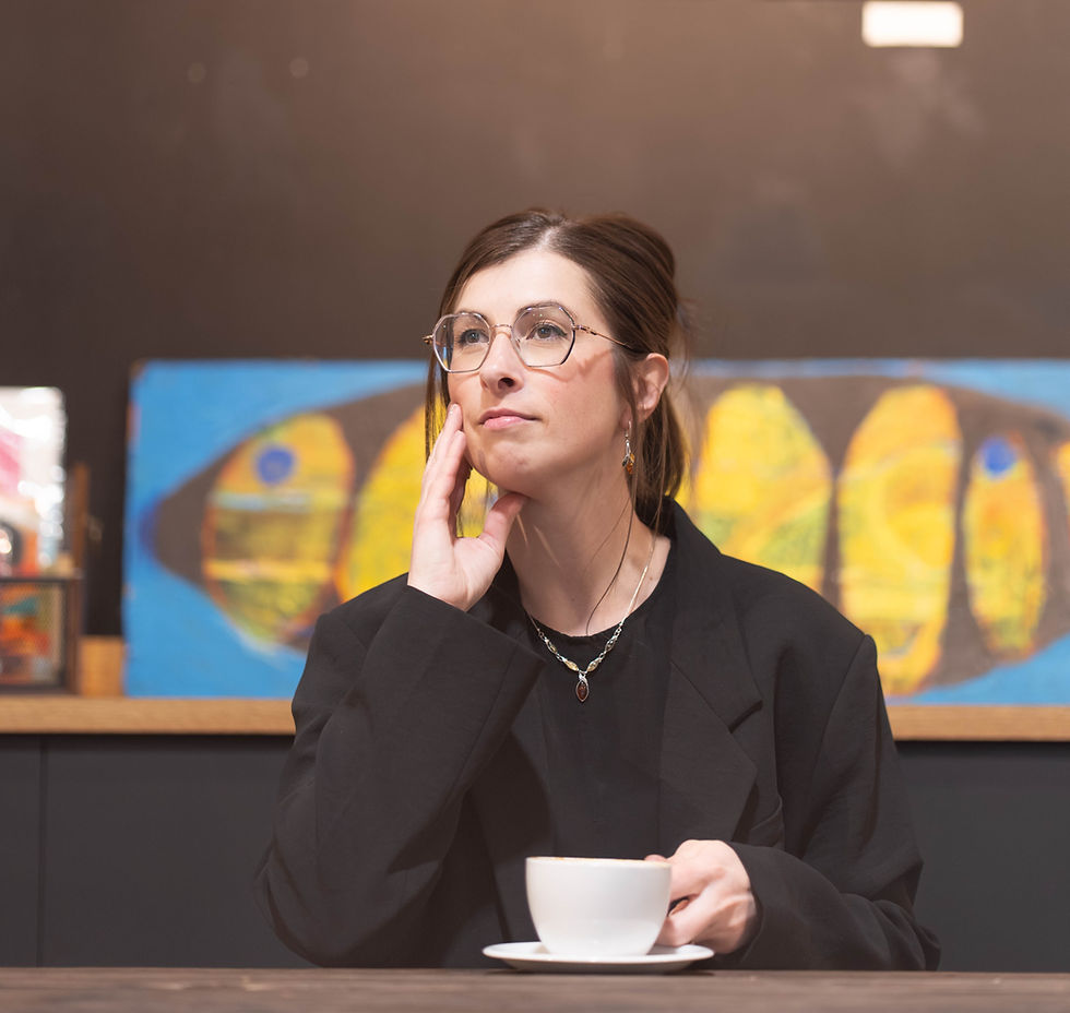 Portrait d’Audrey assise dans un café, regard tourné au loin avec un air contemplatif, lunette et main posée sur son visage. Elle semble réfléchie, comme si elle explorait une question intérieure importante. Une tasse de café chaud est devant elle.