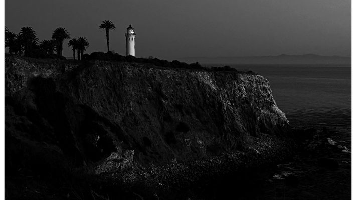 Black and white photo of a solitary lighthouse on a cliff at dusk, evoking stillness, presence, and divine silence.