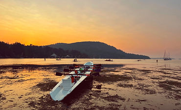 Backgound graphic of an empty coastal quad, oars crossed neatly with blads on the square, resting just off the gunwhales. craft is sitting on its trailer on the wet sand of a flat beach at low tide. the sky is glowing with warmth as the sun is soon to rise.
