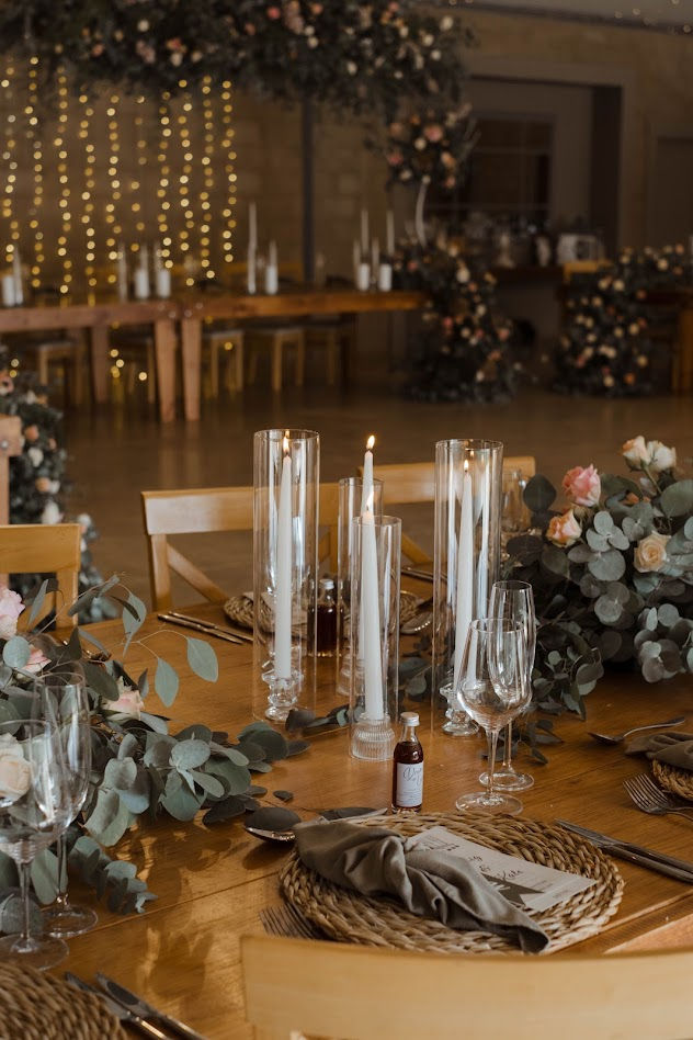 A beautifully set table glows with candlelight, surrounded by elegant floral arrangements and soft greenery, ready for an intimate celebration in a warmly lit venue.