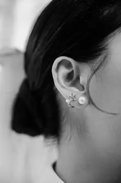 A black-and-white, close-up shot of a woman's ear adorned with elegant jewelry. She wears a large pearl stud alongside a delicate floral-shaped earring. Her dark hair is pulled back into a soft bun, with a few loose strands framing her face.