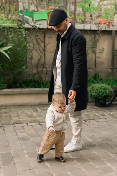 A man in a black flat cap and navy coat holds the hand of a toddler as they walk together across a cobblestone courtyard.