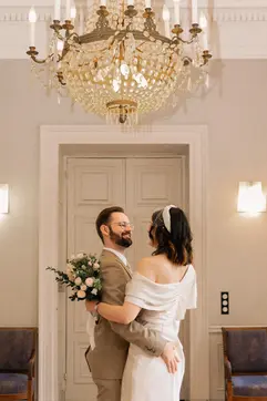 **Romantic couple moment.**

The newlyweds share a tender embrace, smiling at each other under the grand chandelier. She holds her bouquet while he has his arm around her waist. A sweet and intimate wedding portrait.