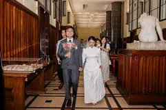 A bride in a white gown and a groom in a grey suit walk hand-in-hand down a grand, wood-paneled hallway, followed by wedding guests. To the side, a marble sculpture and an architectural model sit on pedestals under warm, indoor lighting.