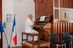 An older man in a white short-sleeved shirt and white trousers plays a large, ornate wooden pipe organ. To his left, two French flags are displayed against a white wall, and a few black chairs sit in the foreground.