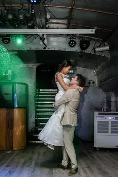 A groom in a beige suit lifts the bride in a white dress, smiling at each other on a dance floor with a green light effect and metallic backdrop. Romantic atmosphere.