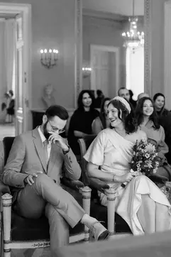 A black and white image of the bride and groom sharing a laugh while seated during their ceremony in an elegant, high-ceilinged hall.