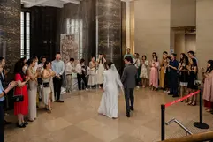 The bride and groom are seen from behind as they walk through a grand lobby with massive dark marble columns. Guests line the sides of the hall, smiling and clapping as the couple passes. The bride wears her white gown and veil, while the groom is in a grey suit, leading the way toward an exit or stairs.