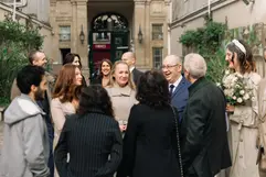 A bride in a birdcage veil and trench coat stands with a group of wedding guests chatting and laughing in a courtyard in Paris.