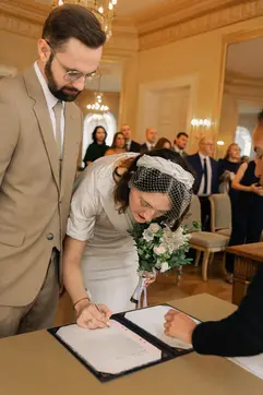 **Wedding signing moment.**

The bride is signing the marriage register while holding her white floral bouquet. The groom stands beside her, watching attentively. They’re in the same elegant hall with guests in the background.