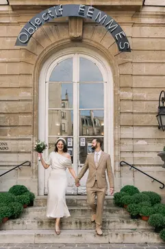 A bride and groom walk hand-in-hand down the stone steps of a building featuring a sign that reads "Objectif FEMMES." The bride, in an elegant off-the-shoulder gown and birdcage veil, smiles at the camera while holding her bouquet, as the groom in his tan suit looks toward her.