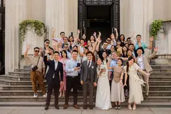 A joyful group photo of a wedding party waving on the wide stone steps of a grand building. The bride and groom stand at the center, surrounded by smiling guests, with tall columns and the words "Liberté Égalité Fraternité" visible above the entrance.