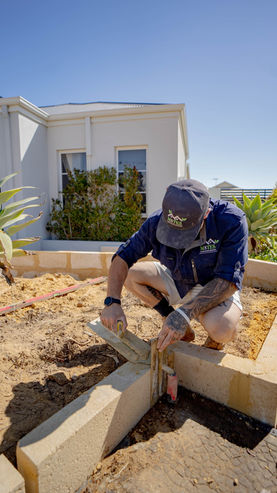 Man laying stone blocks for landscaping, M8tes Landscaping with a house in background.
