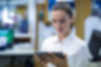 Woman in a white shirt looking at a tablet in a bright, blurred office background. Focused and calm expression.