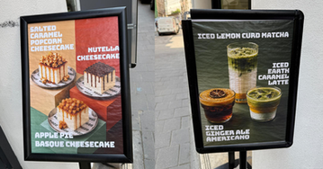 Two sidewalk signs advertise cheesecakes and iced drinks, with colorful dessert and matcha photos on a city street.