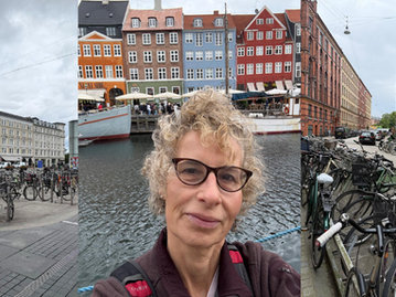 A person with glasses in front of colorful buildings by water, flanked by streets lined with bicycles on a cloudy day.