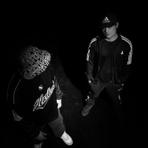 Dos personas con ropa deportiva, una con gorra estampada y sudadera que dice "Harlem", otra con gorra negra. Fondo oscuro, luz de arriba.