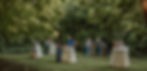 People gather at cocktail tables in a lush, green outdoor setting. Sunlight filters through trees, creating a relaxed, social atmosphere.