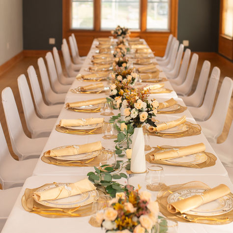 Elegant banquet table set with white chairs, gold plates, cream napkins, and floral centerpieces in a warmly lit room.