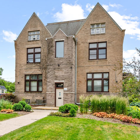Historic brick building with pointed roofs, large windows, and lush gardens. Sky is clear blue. Address "3121" visible on door.