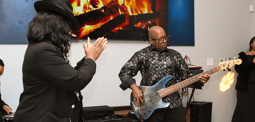 Man plays bass guitar, wearing patterned shirt, beside a woman clapping in black attire. Fireplace video on screen, creating warm ambiance.