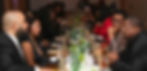 Dinner party with guests seated at a long table, conversing and enjoying drinks. Table adorned with greenery. Semi-formal attire, warm ambiance.