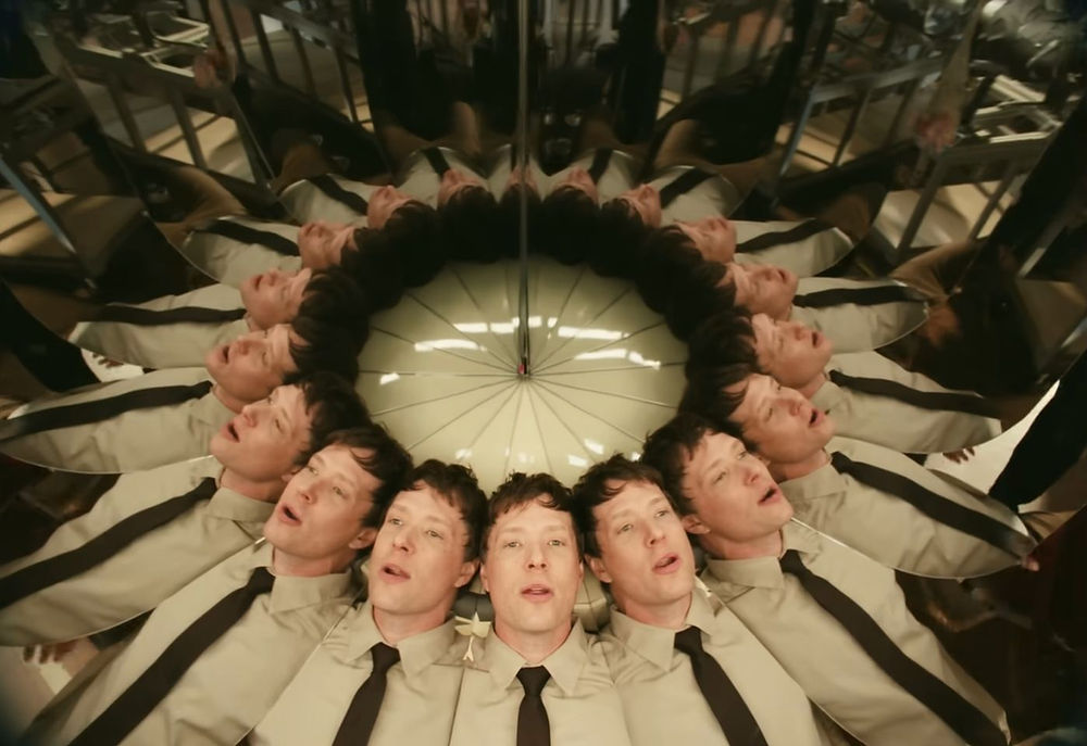 A central performer surrounded by repeated mirrored versions of the same face arranged in a circular pattern, creating optical illusion, self-duplication, and multiplicity.