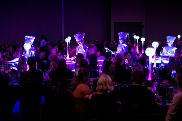 LED lit drummers performing as they enter an awards dinner, with guests seated at tables under purple lighting.