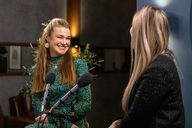 Speaker smiling while talking to a host, with two microphones in the foreground in an indoor recording space.