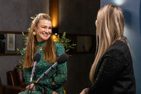 Speaker smiling while talking to a host, with two microphones in the foreground in an indoor recording space.