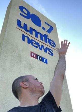 Mark Baratelli, Publisher of The Daily City, stands in front of the WMFE 90.7 Public Radio sign in a half-Evita pose.
