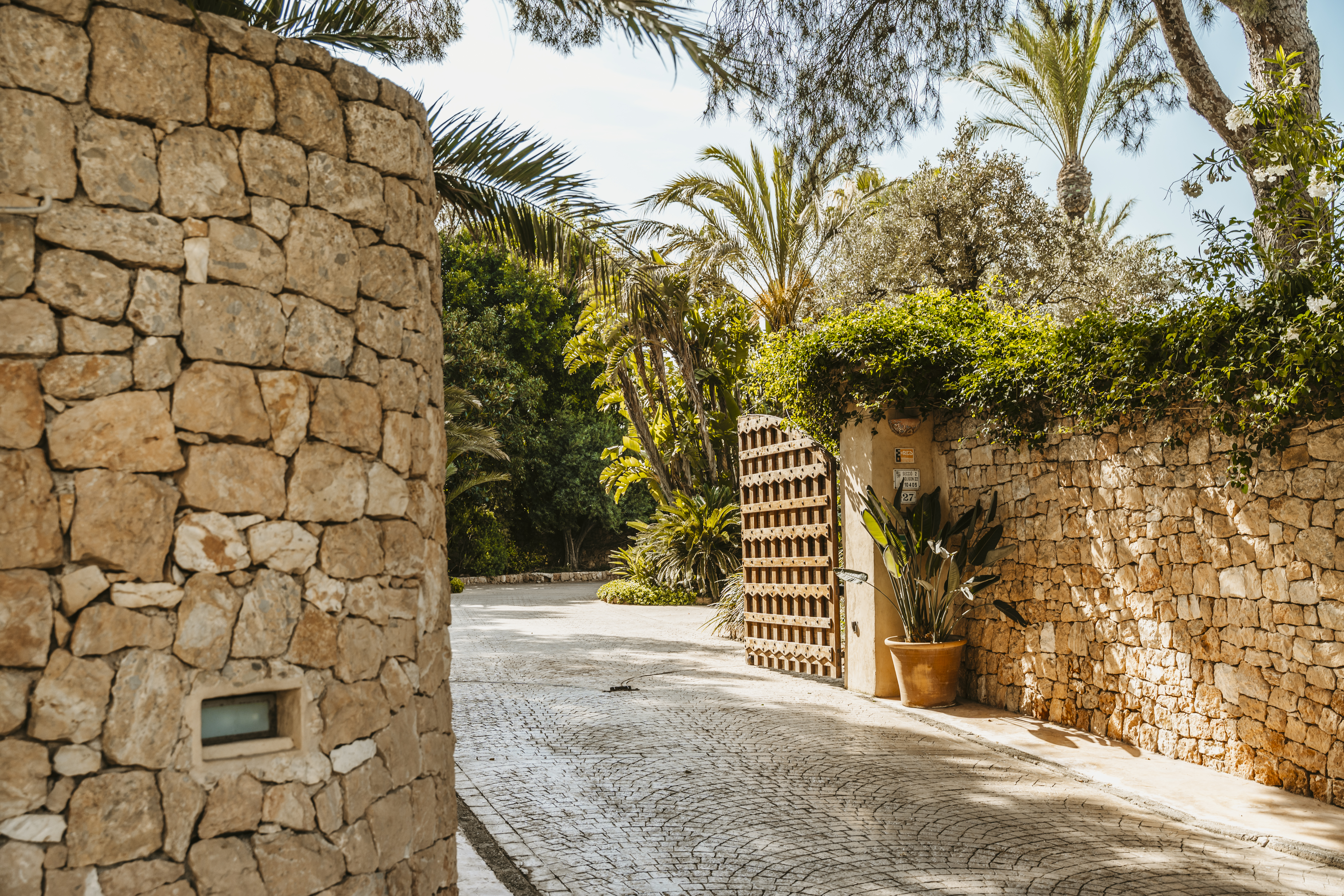 Private entrance of a luxury villa featuring traditional stone walls, a wooden gate, and lush palm trees.