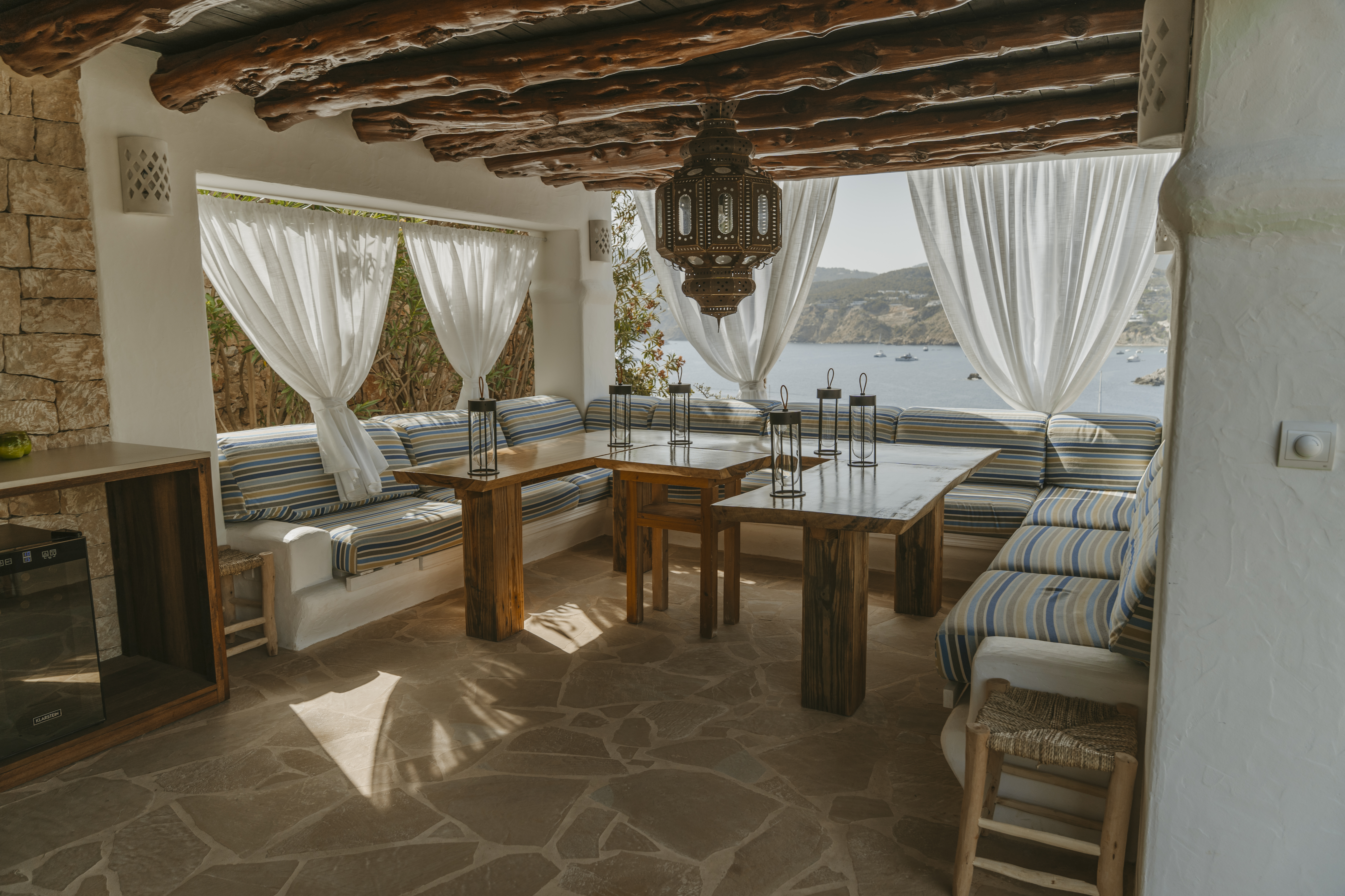 Shaded outdoor dining area with a rustic wooden table, bohemian seating, and panoramic sea views.