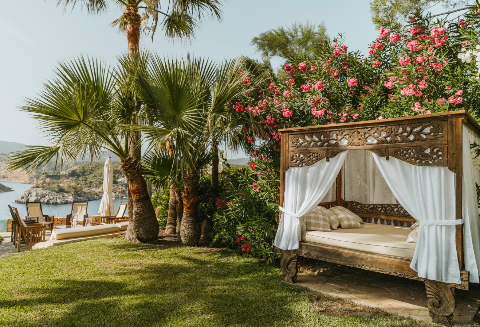 Luxury Balinese daybed on a villa lawn surrounded by pink flowers, palm trees, and coastal views.