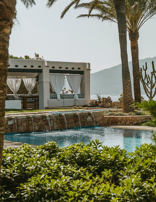 Luxury villa swimming pool with a stone waterfall feature, palm trees, and a white poolside pergola lounge.