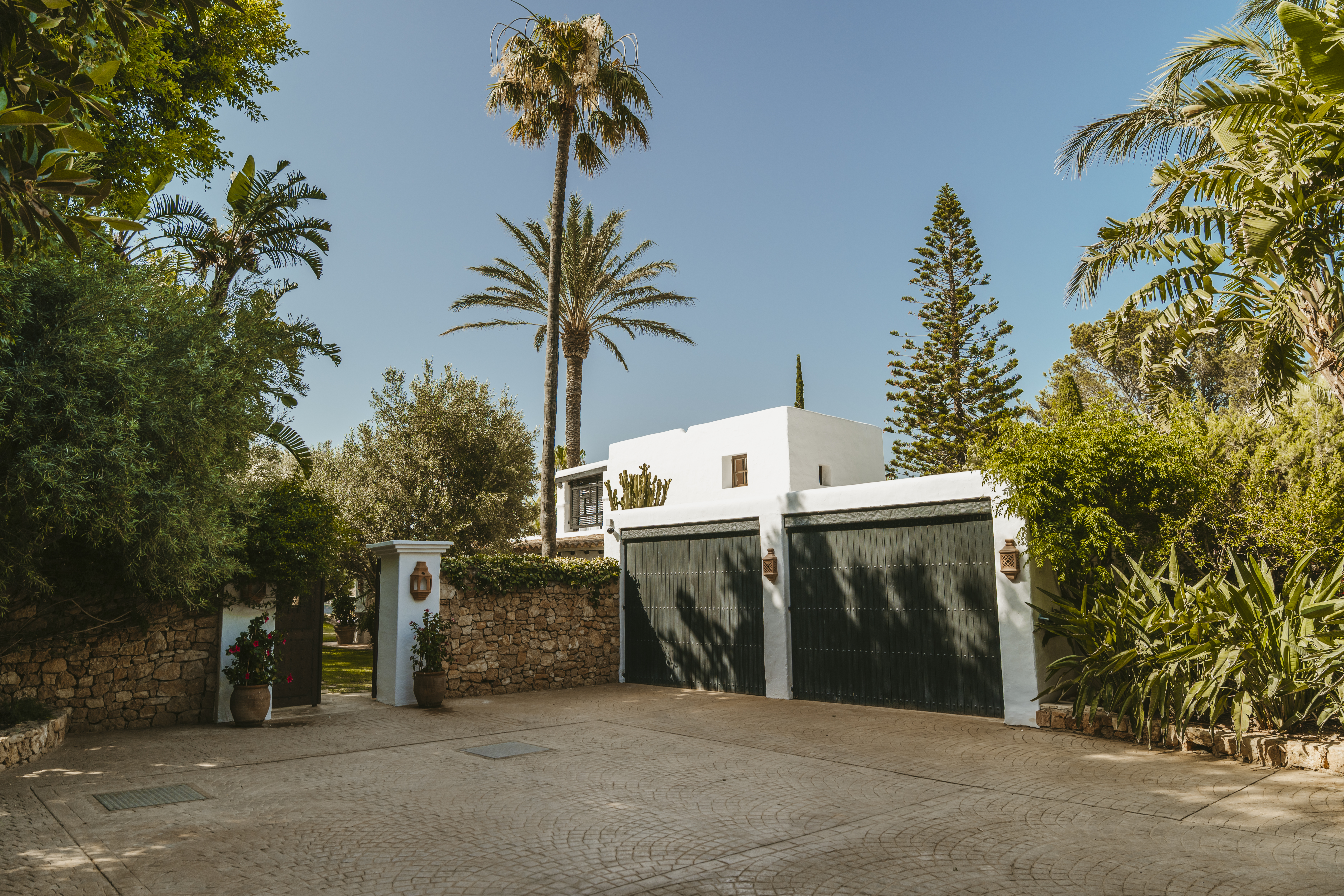 Luxury villa private driveway with a double garage, traditional stone walls, and tall palm trees under a blue sky.