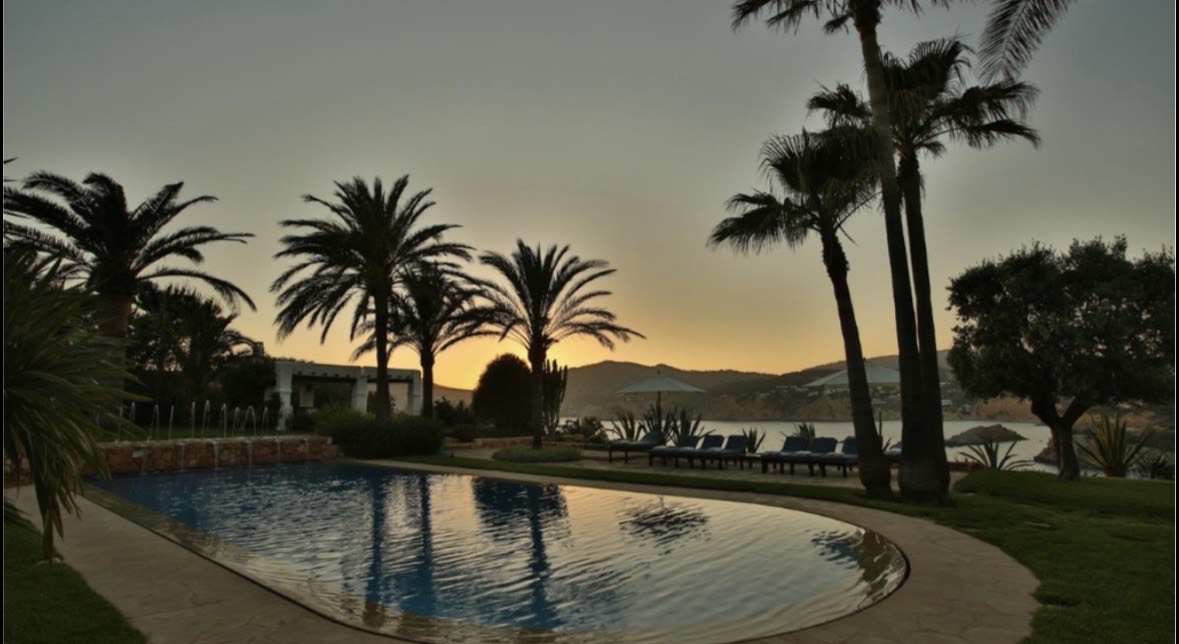 Luxury villa swimming pool at sunset surrounded by tropical palm trees and lounge chairs with coastal views.