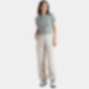 Woman in a sage green top and cream cargo pants stands against a white background, hands in pockets, wearing white boots, looking relaxed.