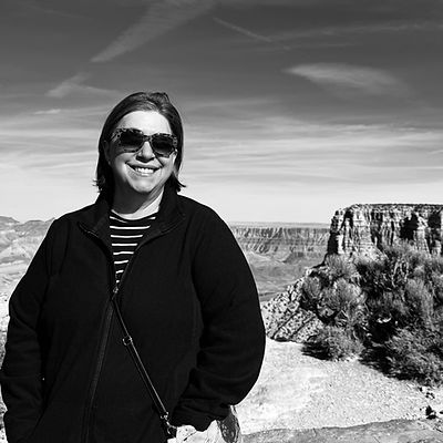 A woman in a black coat and striped shirt, wearing sunglasses, standing before the Grand Canyon in the distance.