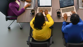 Three people in colorful blazers sitting at a conference table using laptops, with coffee cups and water bottle nearby. Collaborative work setting.