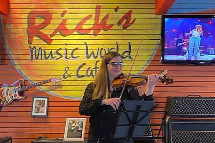Photo of a woman playing violin. She stands in front of a wall that reads "Rick's Music World & Cafe." A guitar hangs on the wall.