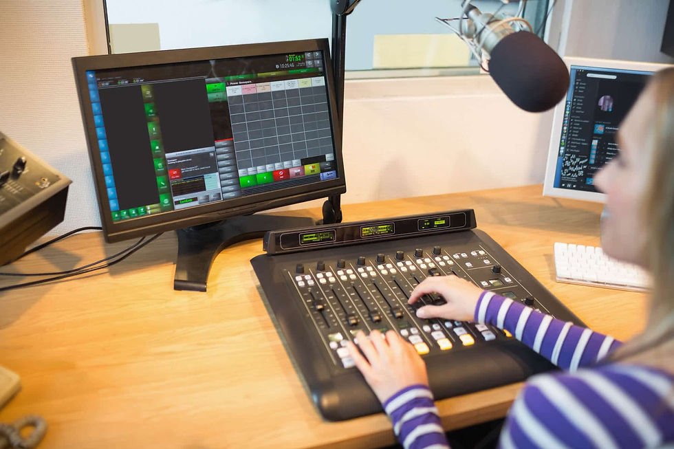 A imagem mostra um estúdio de rádio, onde uma pessoa opera uma mesa de som com vários controles de áudio. À frente, há um monitor exibindo um sistema de programação de rádio, e ao lado um microfone profissional, indicando uma transmissão ou gravação em andamento.