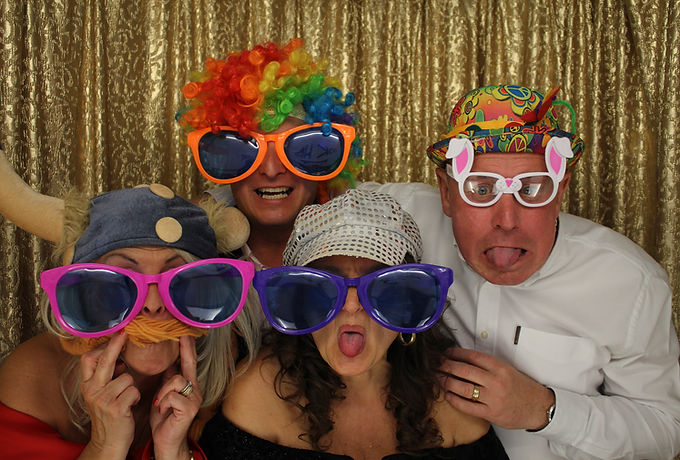 Group of 4 adults in a photo booth, wearing novelty glasses, hats & a wig